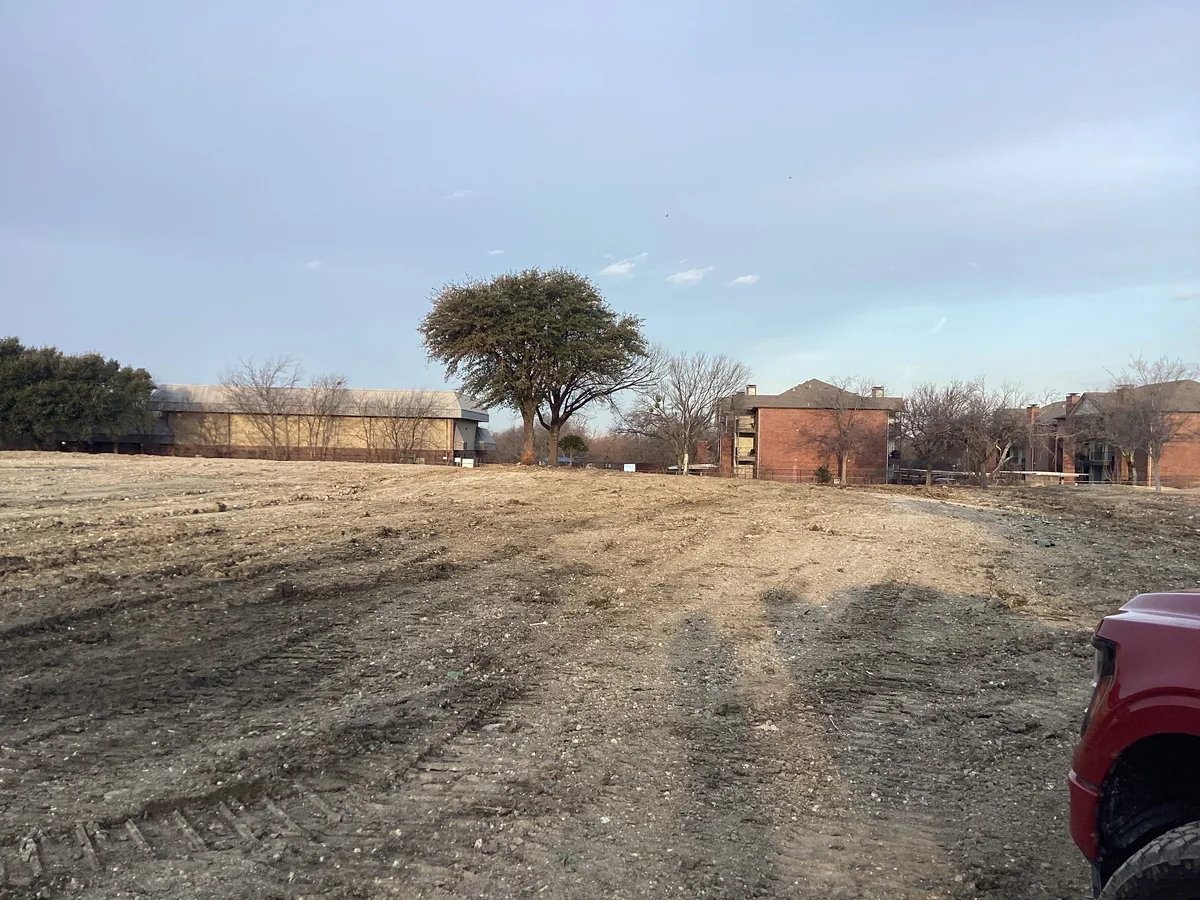 Cleared and graded dirt lot with a single tree and brick buildings