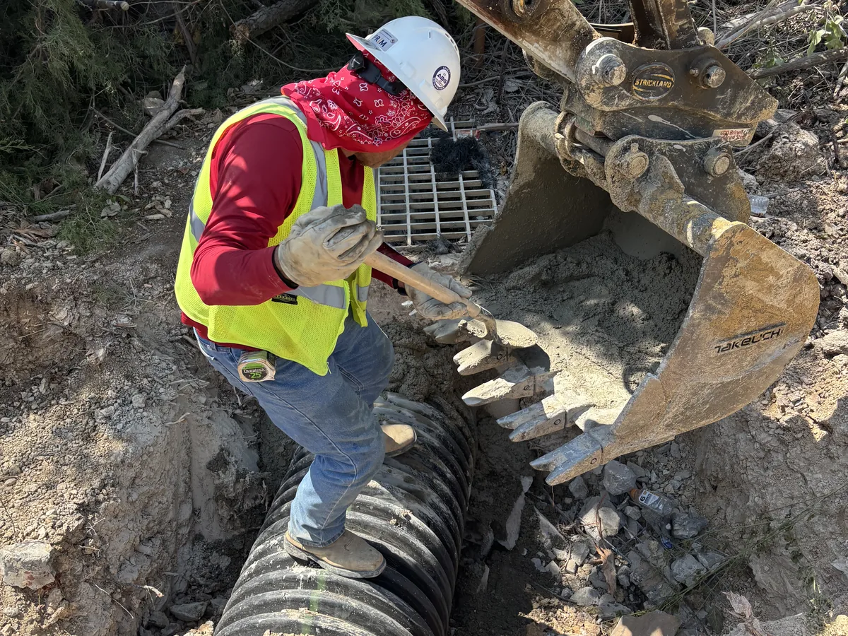 Employee guiding a corrugated storm pipe into place with an excavator bucket