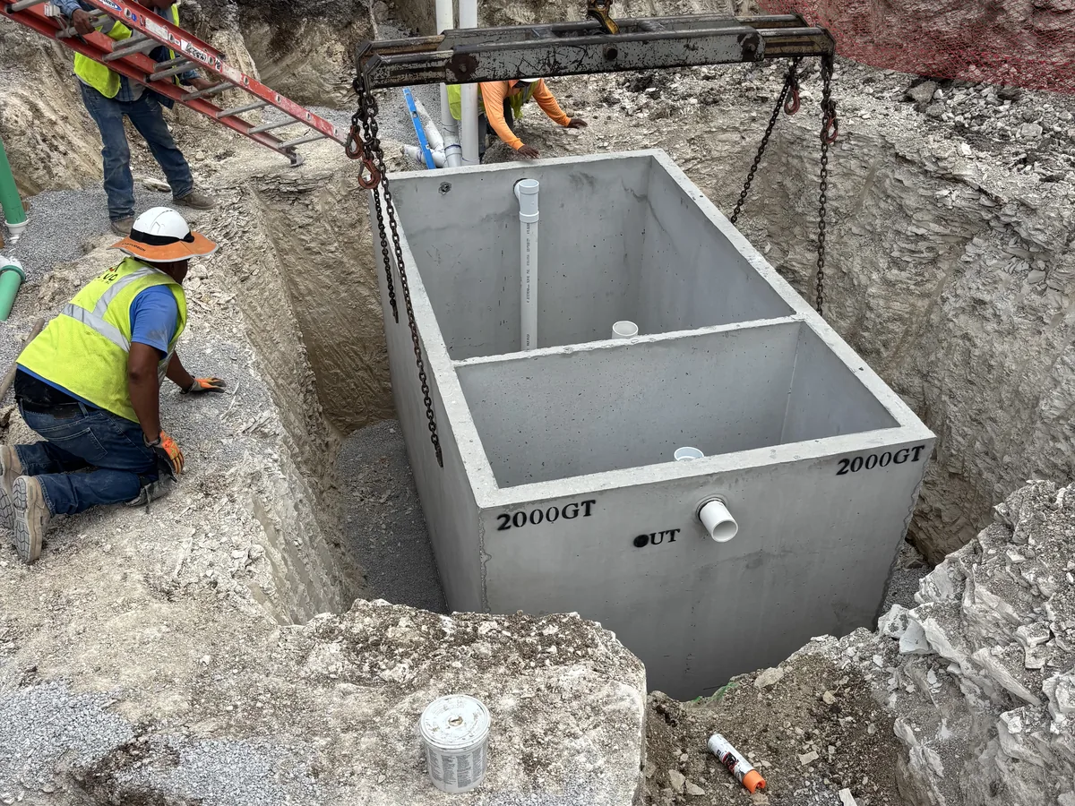 Crew guiding a 2000 gallon precast concrete grease interceptor into an open excavation on crane straps