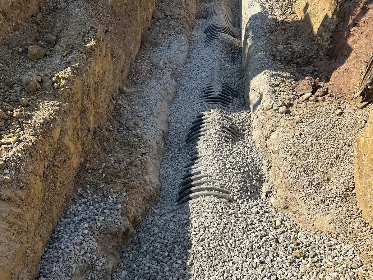 Worker kneeling beside a corrugated pipe set on gravel bedding in a trench