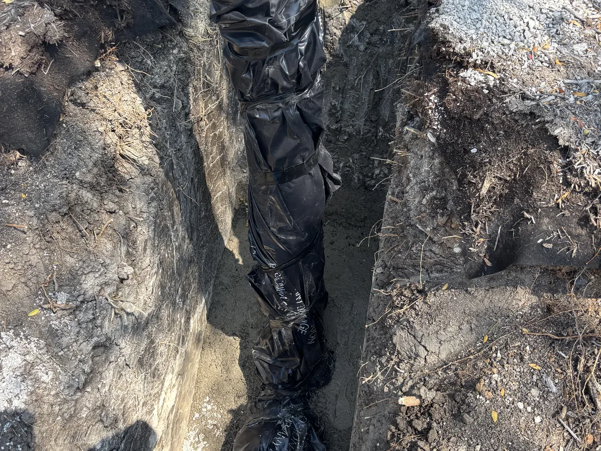 Narrow trench cut through rocky ground with black liner material laid along one wall