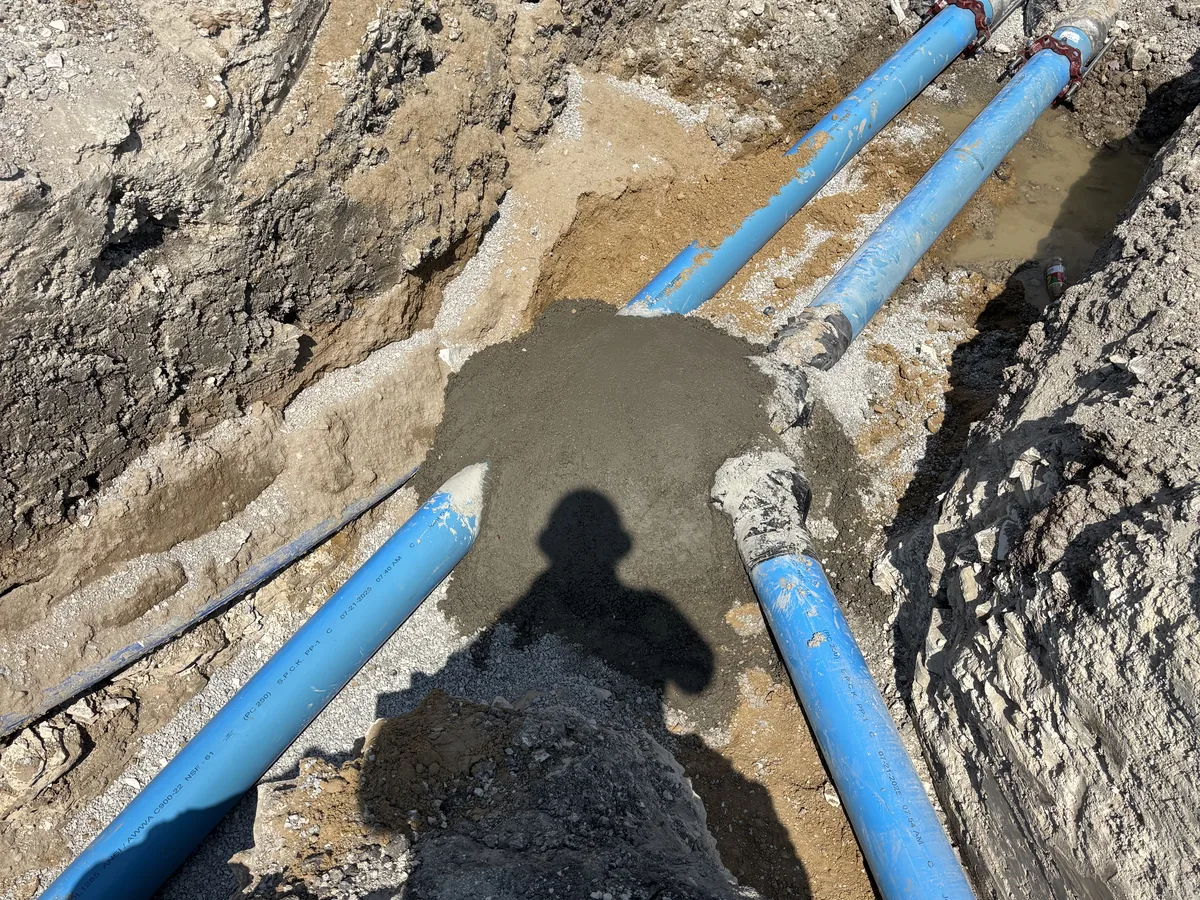 Two blue water lines meeting at a freshly poured concrete thrust block in a trench